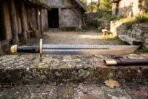 Rune engraved Damascus sword with a broad layered falchion blade carrying an Elder Futhark inscription and gilt brass hilt on a stone ledge in a farm courtyard