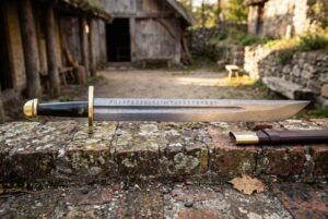 Rune engraved Damascus sword with a broad layered falchion blade carrying an Elder Futhark inscription and gilt brass hilt on a stone ledge in a farm courtyard