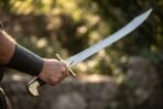 Scimitar with a bright curved blade held out in one hand by its black grip, brass cross guard and hooked pommel, stone wall behind