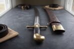 Scimitar with a curved fullered blade, brass cross guard and hooked brass pommel beside a tooled brown leather scabbard with brass chape