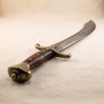Scimitar hilt with brass pommel and riveted wood grip, the curved high carbon blade rising behind it on canvas