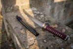 Short Japanese sword with red cord wrapped handle and round guard on a stone ledge beside its black saya