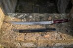 Short Japanese sword with red cord wrapped handle laid along a lichen covered stone wall above its black saya