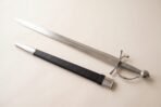 Short sword with a faceted steel pommel, wire bound grip and ring guard laid on a plain white background beside a black scabbard with steel tip