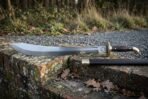 Side view of a Damascus falchion with clipped point and brass hilt resting on a brick wall beside its scabbard