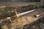 Side view of a dueling sword with gilt brass ring guard and brown wood grip on a sunlit stone ledge
