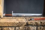 Side view of a high carbon steel falchion with clipped single edged tip propped on a stone wall above its red leather scabbard