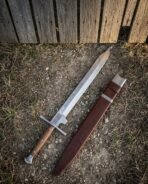 Single edged engraved steel sword with wood grip and steel cross guard lying beside a brown leather scabbard on dry ground