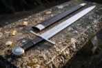 Steel arming sword and black leather scabbard with steel throat lying side by side on a weathered stone slab, low angle view