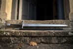 Steel arming sword with black grip and wheel pommel laid beside its black scabbard on a stone doorstep