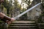Steel arming sword with long fuller and straight cross held out in front of old stone steps