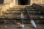 Steel arming sword with wheel pommel standing upright beside its black leather scabbard at the foot of stone stairs