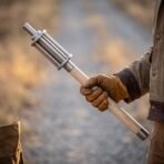 Steel bar mace with pale ash haft and steel end cap held upright in a gloved hand on a country track