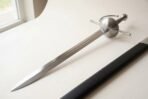 Steel cup hilt rapier with long straight quillons and slender blade on a pale table beside its black scabbard