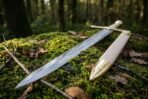 Steel elven longsword blade and cream leather scabbard with brass tip resting together on a mossy log among fallen oak leaves