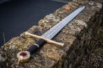 Steel longsword with brass crossguard, black grip and brass disc pommel set with a red stone resting on a mossy stone wall