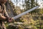 Steel longsword with brass guard and pommel held in both hands by a man outdoors, blade raised at an angle in low sun