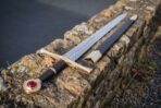 Steel longsword with brass hilt and red stone pommel lying on a stone wall beside its black scabbard with brass fittings