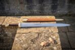 Steel longsword with fullered blade and engraved brass hilt laid on a stone ledge with the tan leather scabbard behind it