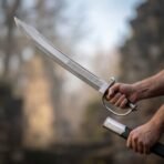 Steel naval cutlass with knotwork etched blade and stirrup guard drawn from a black scabbard held in the other hand outdoors