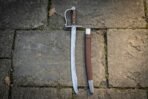 Steel pirate cutlass with wooden grip and knuckle bow standing upright beside its brown leather scabbard on stone paving