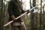 Steel poleaxe with crescent blade and spike held across the body in gloved hands by a man in a dark wool tunic in a forest