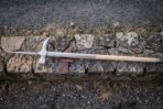 Steel polehammer with hammer face, curved beak and top spike on a long pale ash haft laid along a stone wall with leather cover