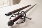 Steel rapier, close up of the blackened swept hilt and pierced pommel set with red stones beside the tooled scabbard