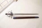 Steel rapier with blackened swept hilt and oxblood grip laid on cream paper above its tooled brown leather scabbard