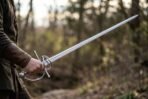 Steel rapier with side ring guard and ball pommel held out at the side on a woodland path