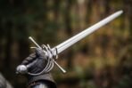 Steel rapier with swept hilt and long thrusting blade held up in a gloved hand against an autumn forest background