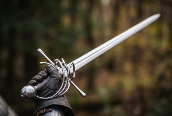 Steel rapier with swept hilt and long thrusting blade held up in a gloved hand against an autumn forest background