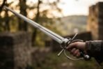 Steel renaissance side sword held out at full stretch by its ring hilt above blurred castle ruins at golden hour