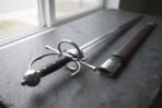 Steel renaissance side sword with ring guard, knuckle bow and ball pommel on a slate window sill beside its brown leather scabbard