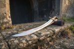 Steel shamshir lying blade forward on a stone sill before an arched doorway, its brown scabbard and a rolled cloth behind