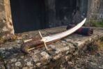 Steel shamshir with hand carved vine scroll grip and brass quillons resting on its scabbard on a lichen covered stone step