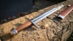 Straight backed high carbon steel falchion with wooden grip and steel cross guard on a sunlit stone ledge by its red scabbard