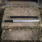 Straight steel dao sword with black cord wrapped grip laid below its black scabbard on a lichen covered stone step