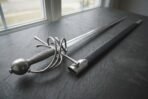 Swept hilt rapier with coiled steel guard and ball pommel seen from the hilt end on dark slate next to its black scabbard