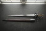 Swept hilt rapier with a long Damascus blade laid flat on dark slate above its brown leather scabbard, seen from above