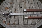 Side sword with a swept hilt and long straight blade laid flat on a weathered wooden table, brown leather scabbard with steel fittings above