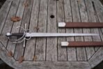 Top view of a side sword with swept steel guards and a round pommel on a wooden table, brown leather scabbard in two sections beside it