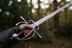 Side sword with a swept steel hilt held in a gloved hand, round pommel and dark grip in focus, blade pointing up into the trees