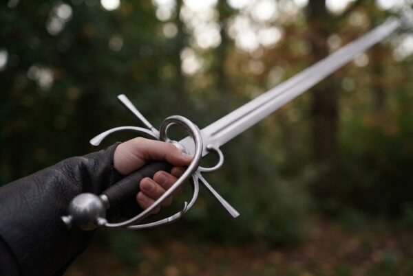 Side sword with a swept steel hilt held in a gloved hand, round pommel and dark grip in focus, blade pointing up into the trees