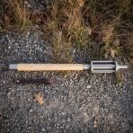 Top down view of a steel bar mace with ash haft laid straight on gravel above its brown leather wrist strap and an oak leaf