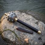 Top view of a Damascus spiked mace head, brass collars and dark haft lying on a wet stone beside a leather wrist strap
