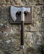 Viking axe with wall mount hung on a stone wall, the hand forged runic bearded axe cradled on a carved timber rune plaque with blackened steel hooks.