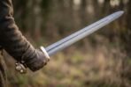 Viking sword blade with a fuller held in a gloved hand, brass guard and lobed pommel visible against a soft autumn field