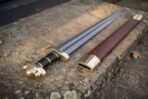 Viking sword with a brass lobed pommel and black grip laid beside its brass tipped brown leather scabbard on a weathered stone step