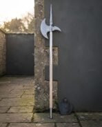Whole Damascus halberd with long steel langets and pale wooden haft standing upright against a grey courtyard wall